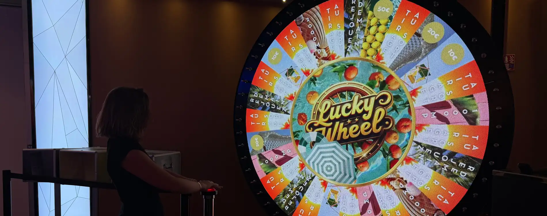 Una donna preme un pulsante rosso per attivare la Lucky Wheel di giugno al Casinò Café de Paris. Dietro di lei si vedono la ruota e tre caselle destinate all’estrazione dei premi per i soci Silver, Gold e Platinum.